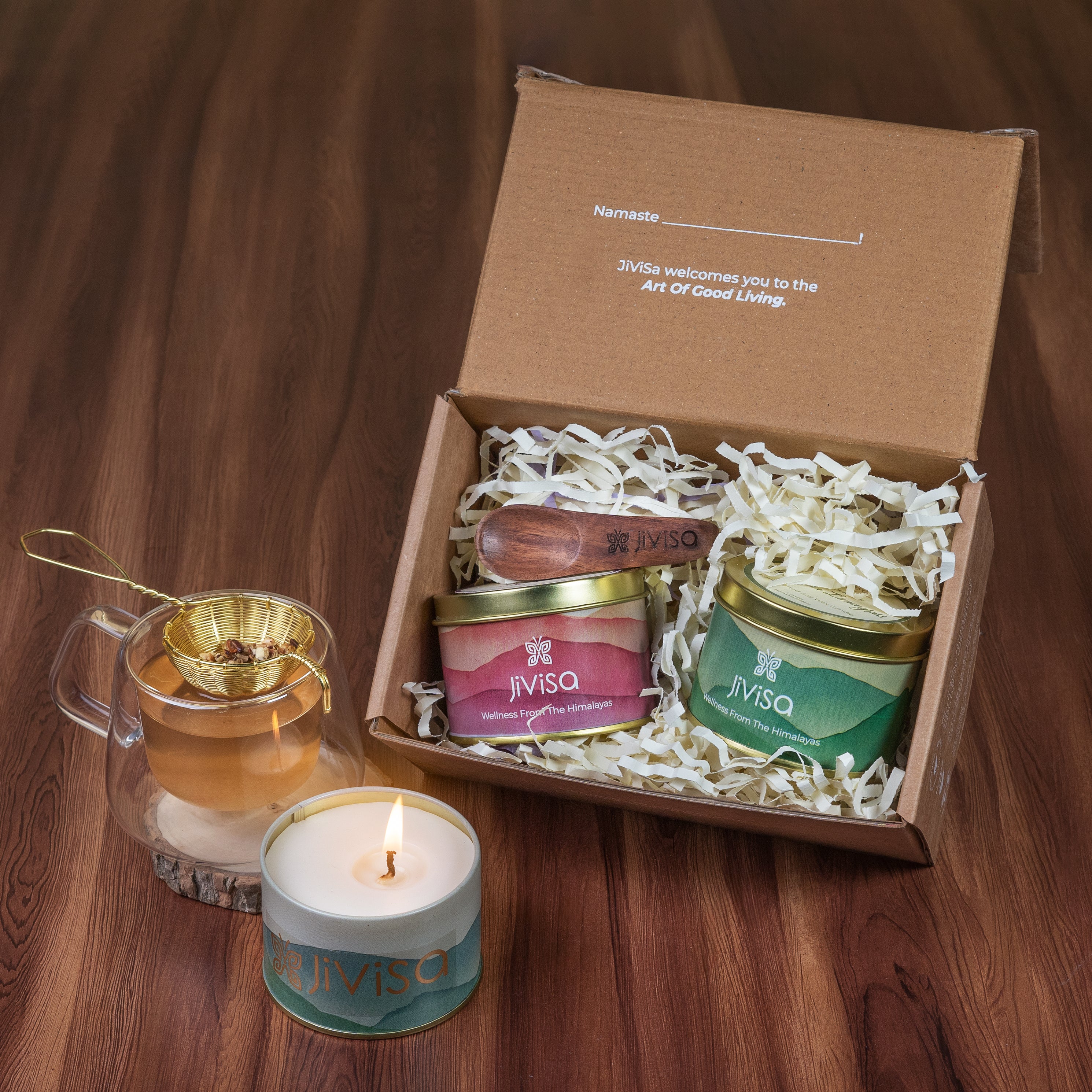 Tranquil Himalayan Collection Gift Box - JiViSa. 
The image shows a gift box containing two jars, one is JiViSa Herbal Tea and other JiViSa Wax candle with a wooden spoon. The box placed on a wooden table alongside another candle in a glass holder and Herbal Tea glass cup.