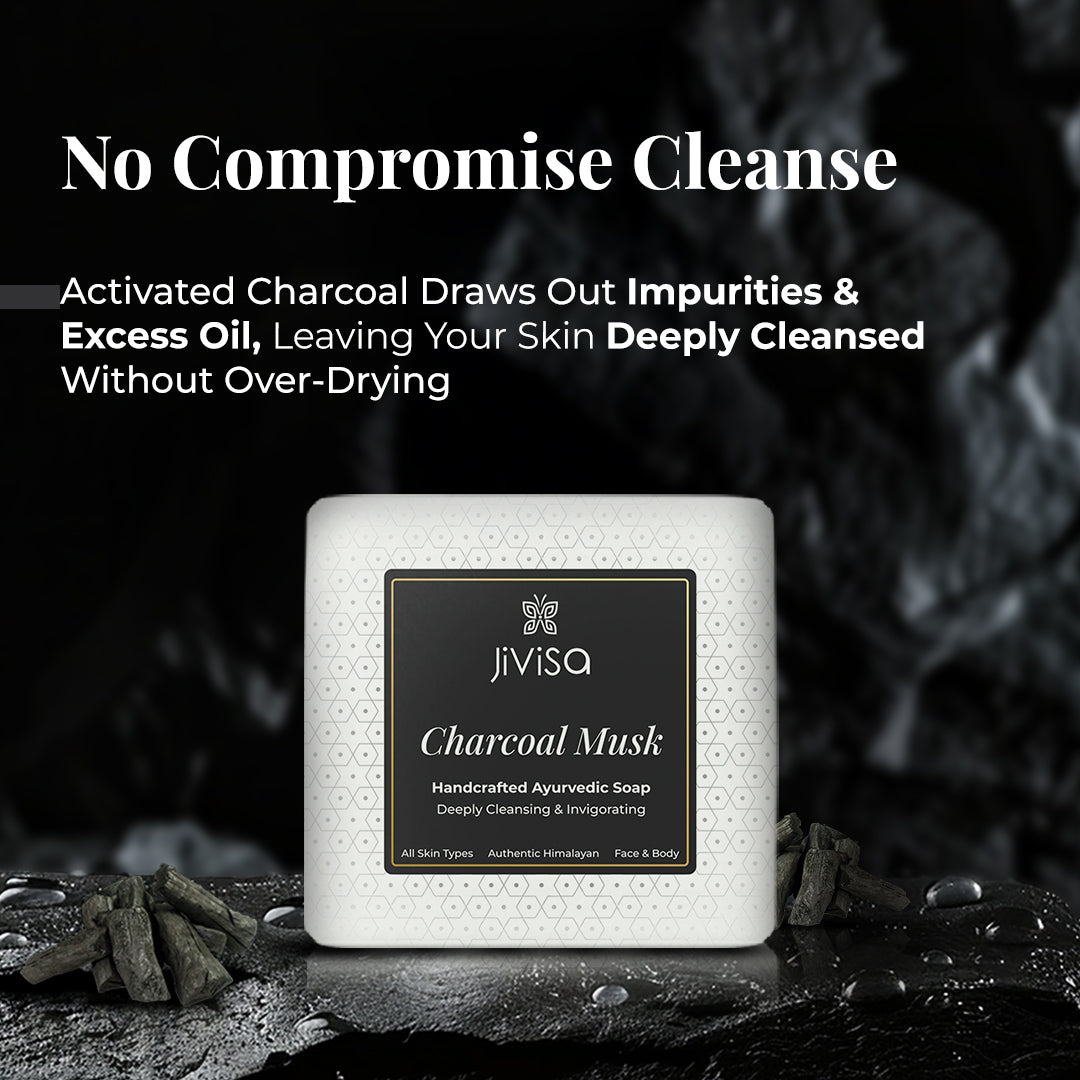 JiViSa Charcoal Musk Handcrafted Ayurvedic Soap bar, in white packaging with a black and white patterned label, displayed on a dark, textured surface with pieces of charcoal around it. The background is dark and gritty. Tagline: "No Compromise Cleanse". Text describe its benefits: Activated Charcoal Draws Out Impurities & Excess Oil, Leaving Your Skin Deeply Cleansed Without Over-Drying.