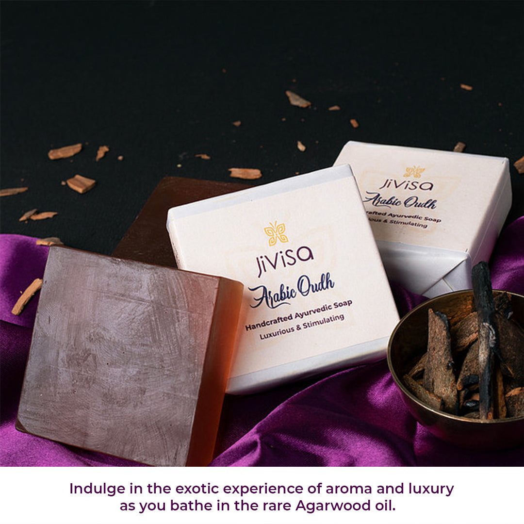 JiViSa Arabic Oudh Handcrafted Ayurvedic Soap captured in a luxurious setting with dark wood tones, rich purple silk, and natural agarwood (Oudh) pieces in a copper bowl. An unwrapped soap bar with a deep brown translucent tone is also displayed, emphasizing its natural composition. Below the setup, the text reads: “Indulge in the exotic experience of aroma and luxury as you bath in the rare Agarwood Oil".