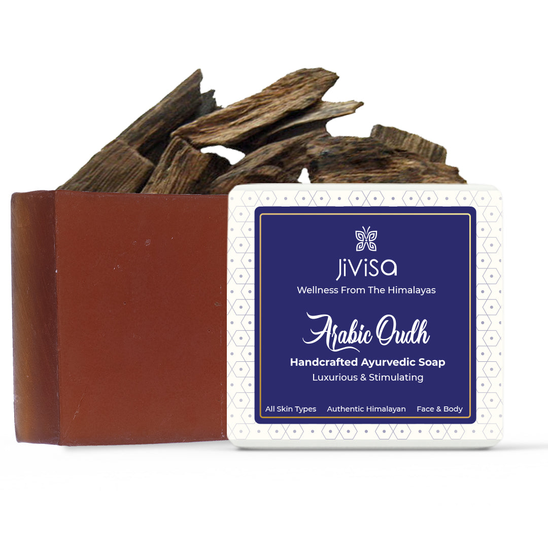 A deep brown JiViSa Arabic Oudh Handcrafted Ayurvedic Soap bar is placed beside its packaging box with a blue and white patterned label, displayed in front of agarwood pieces on the top. The label reads: JiViSa – Wellness From The Himalayas-Arabic Oudh Handcrafted Ayurvedic Soap, Luxurious & Stimulating with notes: &