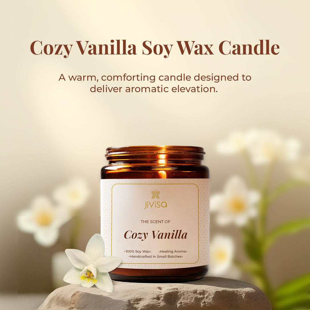 JiViSa Cozy Vanilla candle placed on a stone surface with white vanilla flowers in the background. The tagline above reads: "Cozy Vanilla Soy Wax Candle – A warm, comforting candle designed to deliver aromatic elevation." The label on the jar reads: "The Scent of Cozy Vanilla – 100% Soy Wax • Healing Aroma • Handcrafted In Small Batches."