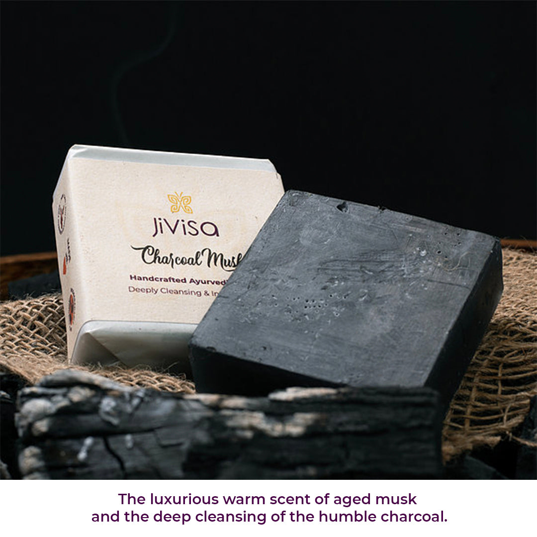 An unwrapped JiViSa Charcoal Musk Handcrafted Ayurvedic Soap bar resting next to its white packaging on a piece of burlap, surrounded by charred coal pieces. A faint wisp of smoke is visible in the background. Bottom text: &