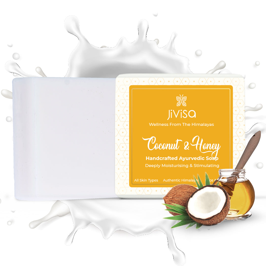 JiViSa Coconut & Honey Ayurvedic Soap Bar next to its white and yellow packaged box. Behind them is a large white splash effect of milk and in the foreground are a whole coconut and a halved coconut, a honey dipper, and a small jar of honey.