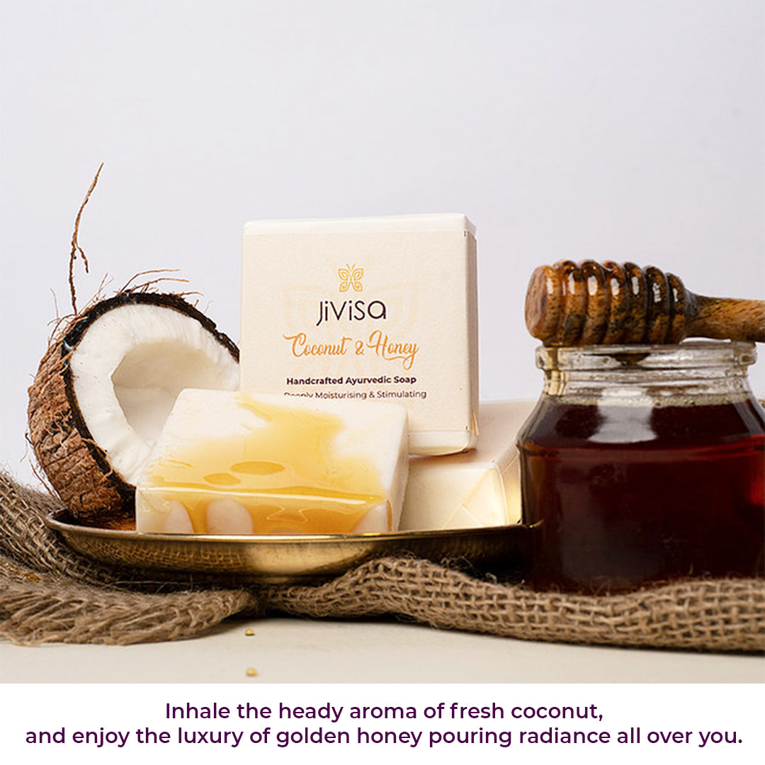 JiViSa Coconut & Honey Handcrafted Ayurvedic Soap bar placed next to its white and gold packaged box. A broken coconut half, a glass jar of honey with a dipper are arranged around it on a textured surface. Bottom text reads: Inhale the heady aroma of fresh coconut and enjoy the luxury of golden honey pouring radiance all over you.