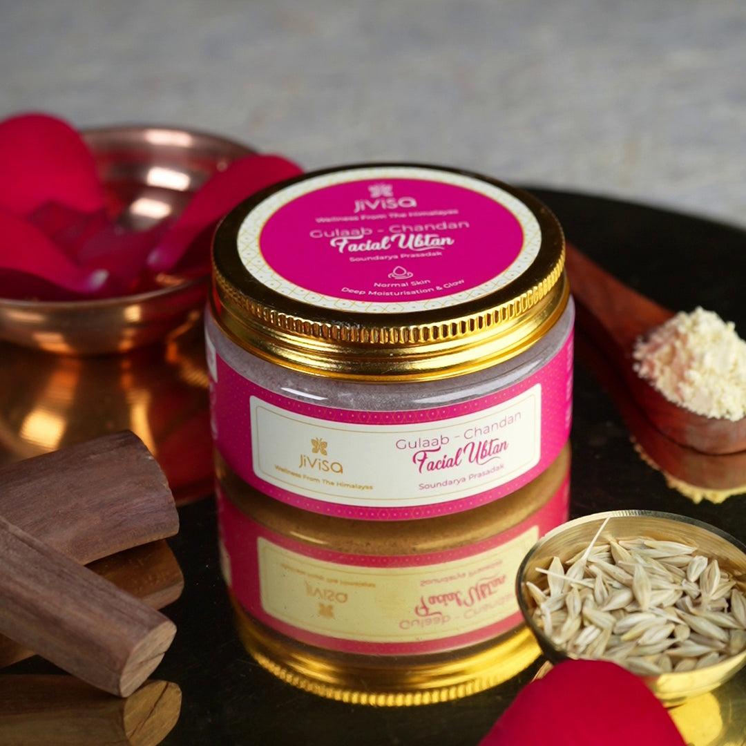 JiViSa Gulaab Chandan Facial Ubtan is the central focus surrounded by various natural ingredients, including sandalwood pieces, a small wooden spoon with a mound of Gram flour powder and a small gold bowl filled with seeds and soft red rose petals are visible in the blurred background.