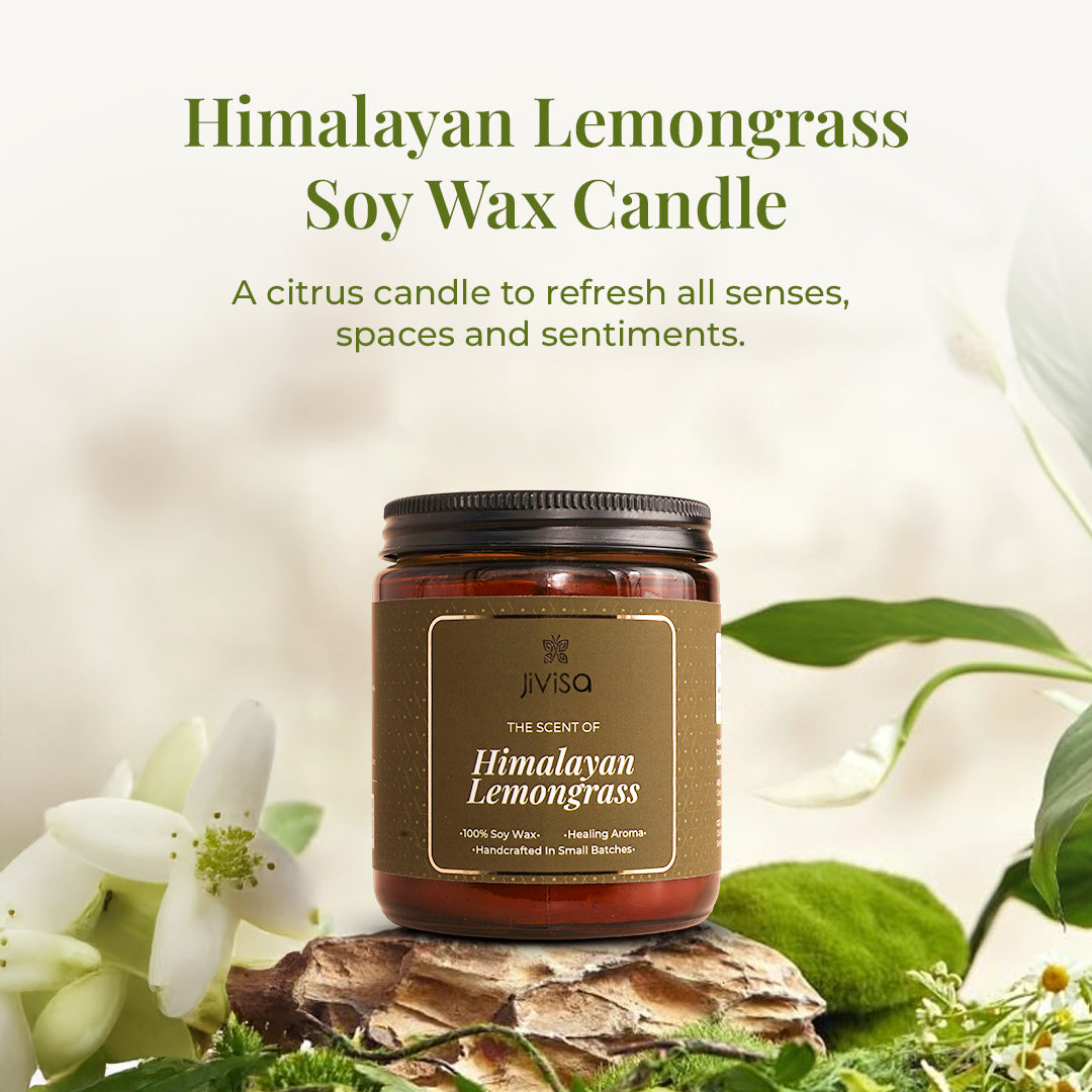JiViSa Himalayan Lemongrass soy wax candle amber glass jar is displayed on a pinecone base surrounded by white flowers and green foliage, with a soft, airy background. The text above reads: "Himalayan Lemongrass Soy Wax Candle – A citrus candle to refresh all senses, spaces and sentiments."