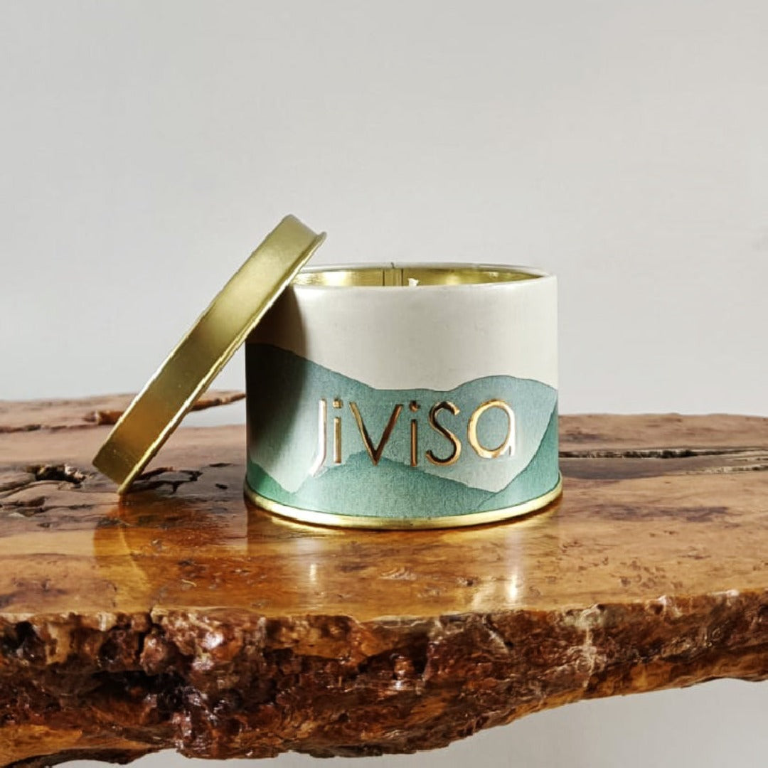 JiViSa Himalayan Lemongrass soy wax candle in a gold metal jar with mountain-print label, placed on a rustic wooden slab with lid slightly open.