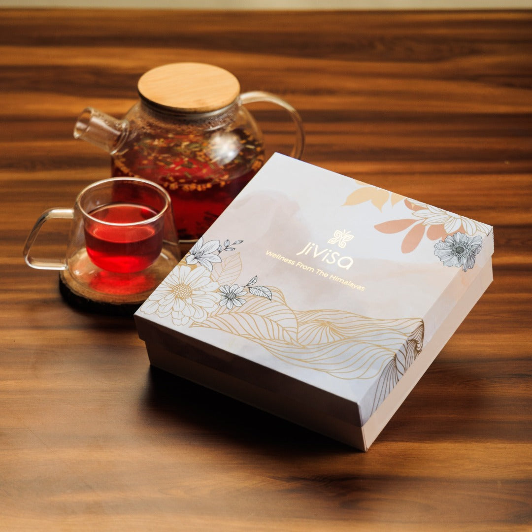 JiViSa Himalayan Discovery Tea Gift Box featuring the tagline "Wellness From The Himalayas" on the lid. Below the gift box, clear glass tea pot and a cup are positioned  with read brown herbal tea.