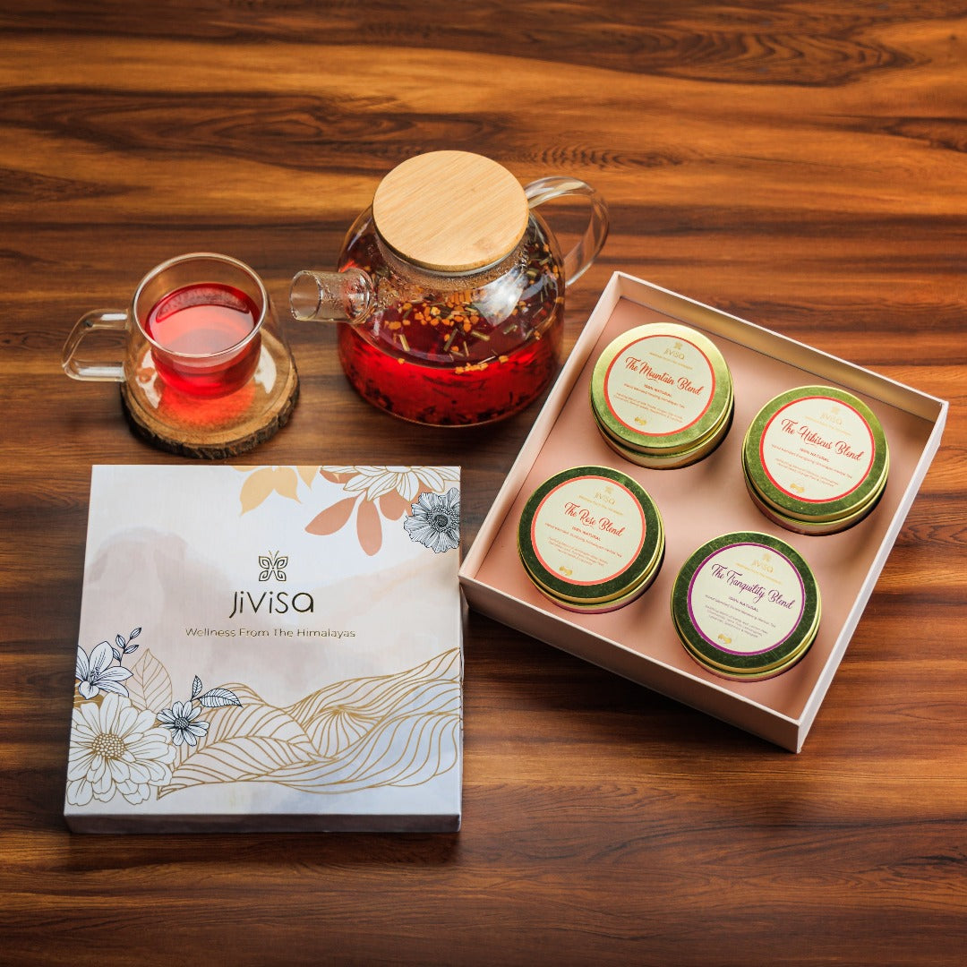 JiViSa Himalayan Discovery Tea Gift Box. The image displays a decorated white box with the JiViSa logo and Wellness From The Himalayas tagline sits on the left. To the right, an open gift box reveals four mini tins of JiViSa Herbal teas : including The Mountain Blend, The Slumber Blend, The Rose Blend, and The Tranquility Blend. In the upper left, a clear glass teapot with a wooden lid and a matching teacup filled with a red-hued tea are visible, the teacup resting on a small wooden coaster.
