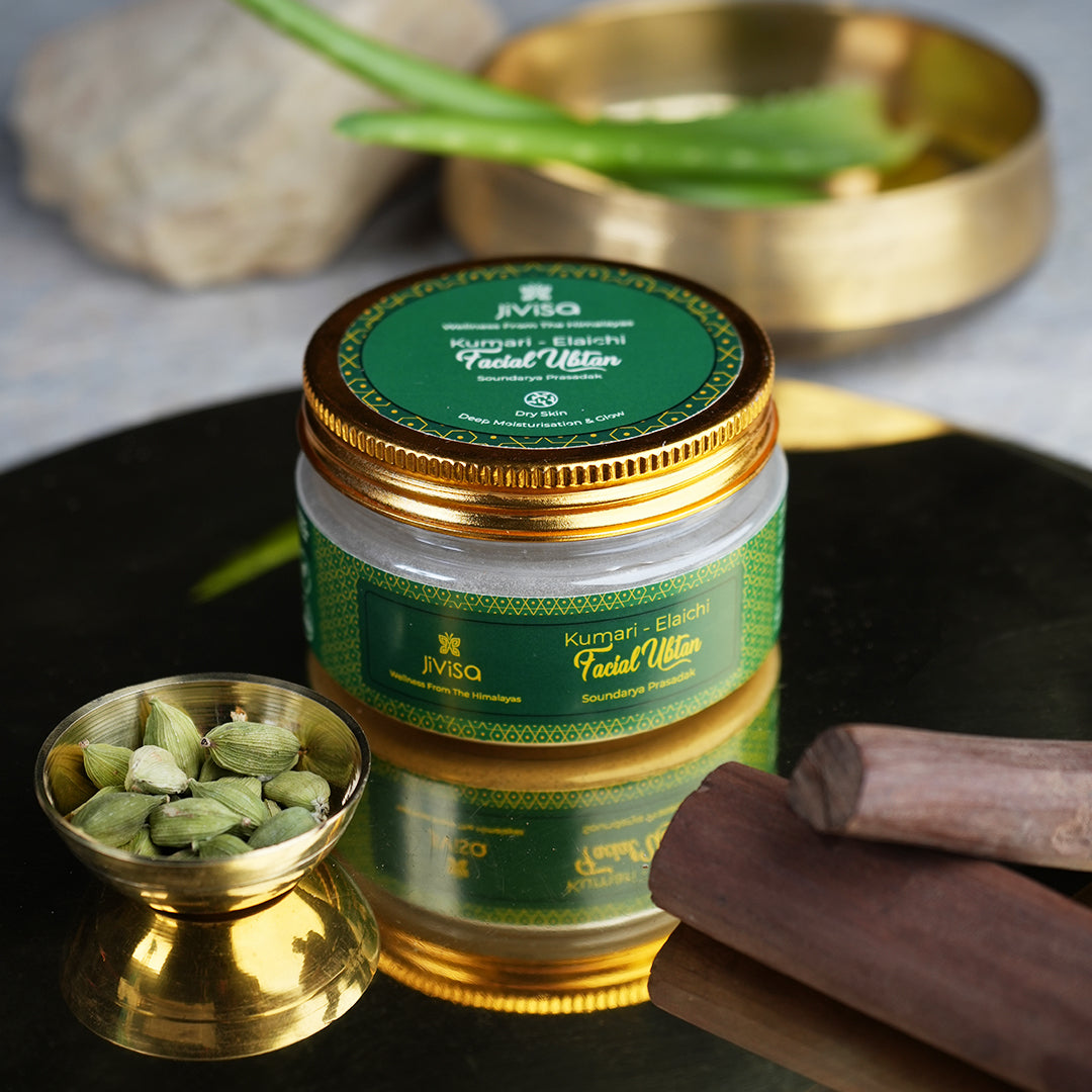 JiViSa-Kumari Elaichi Facial Ubtan is the central focus surrounded by various natural ingredients, including a small gold color bowl of Elaichi, sandalwood, and a BIG bowl contain two Aloevera visible in the blurred background.