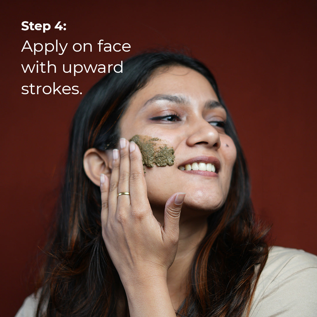A woman with medium brown skin smiles gently as she applies a light purple scrub to her cheek with upward strokes. She is wearing a simple gold ring. Text overlay reads "Step 4: Apply on face with upward strokes.