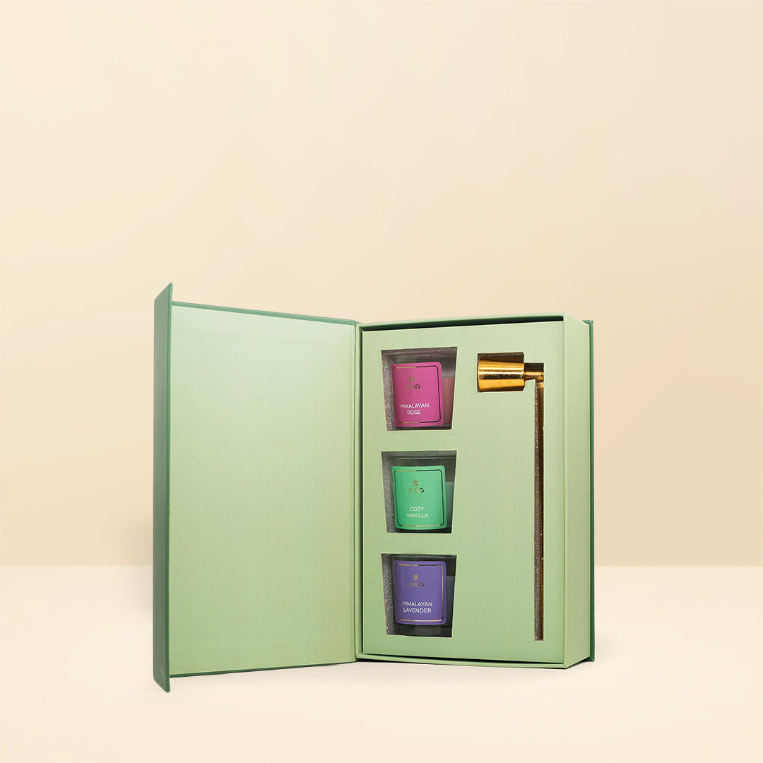 JiViSa Lotus Light Votive Candle Snuffer Gift Box shaped like a book, with a pale green cover. The box is open, revealing three colorful JiViSa votive candles (pink, green, and purple) and a slender golden candle snuffer. The items are arranged neatly in a fitted, pale green insert.