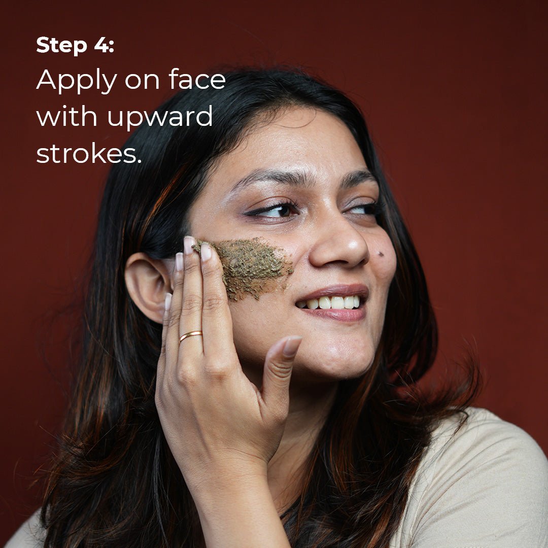A woman with medium brown skin smiles gently as she applies a light purple scrub to her cheek with upward strokes. She is wearing a simple gold ring. Text overlay reads "Step 4: Apply on face with upward strokes