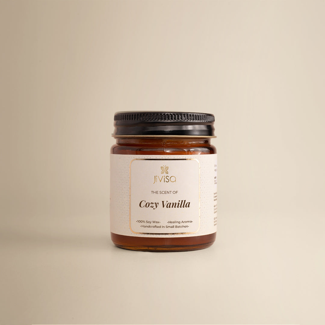 JiViSa Cozy Vanilla soy wax candle in a glossy amber glass jar 90gm with a black lid, placed against a beige background. The label reads: "JiViSa β Wellness From The Himalayas β The Scent of Cozy Vanilla β Handcrafted β’ 100% Soy Wax β’ Healing Aroma."
