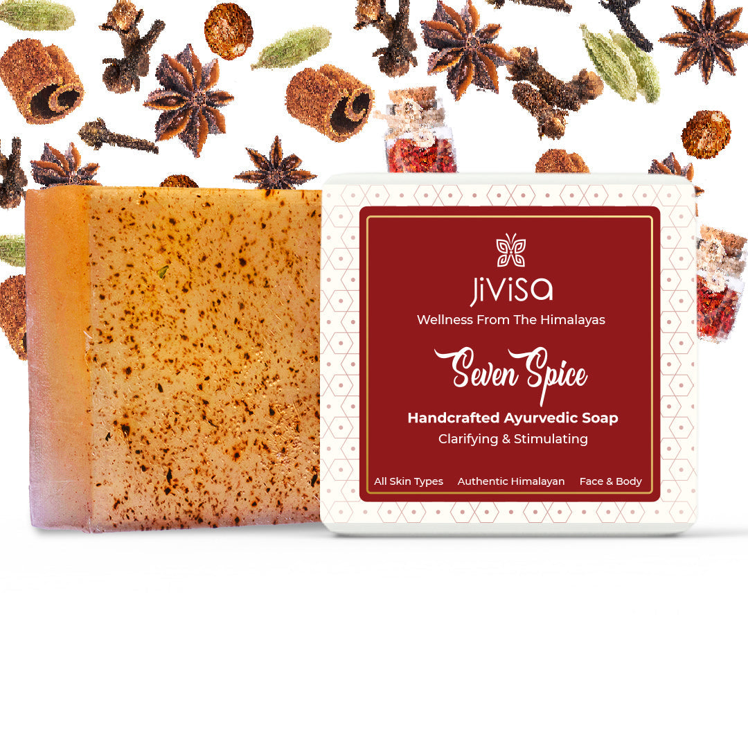 An unwrapped bar of JiViSa Seven Spice Handcrafted Ayurvedic Soap, light brown with dark specks, next to its white and maroon packaged box. The background is a white surface scattered with various whole spices including star anise, cloves, cinnamon sticks, and cardamom pods, appears to be dried spices. Text on box: JiViSa Wellness from the Himalayas Seven Spice Handcrafted Ayurvedic Soap Clarifying & Stimulating All Skin Types-Authentic Himalayan-Face & Body.