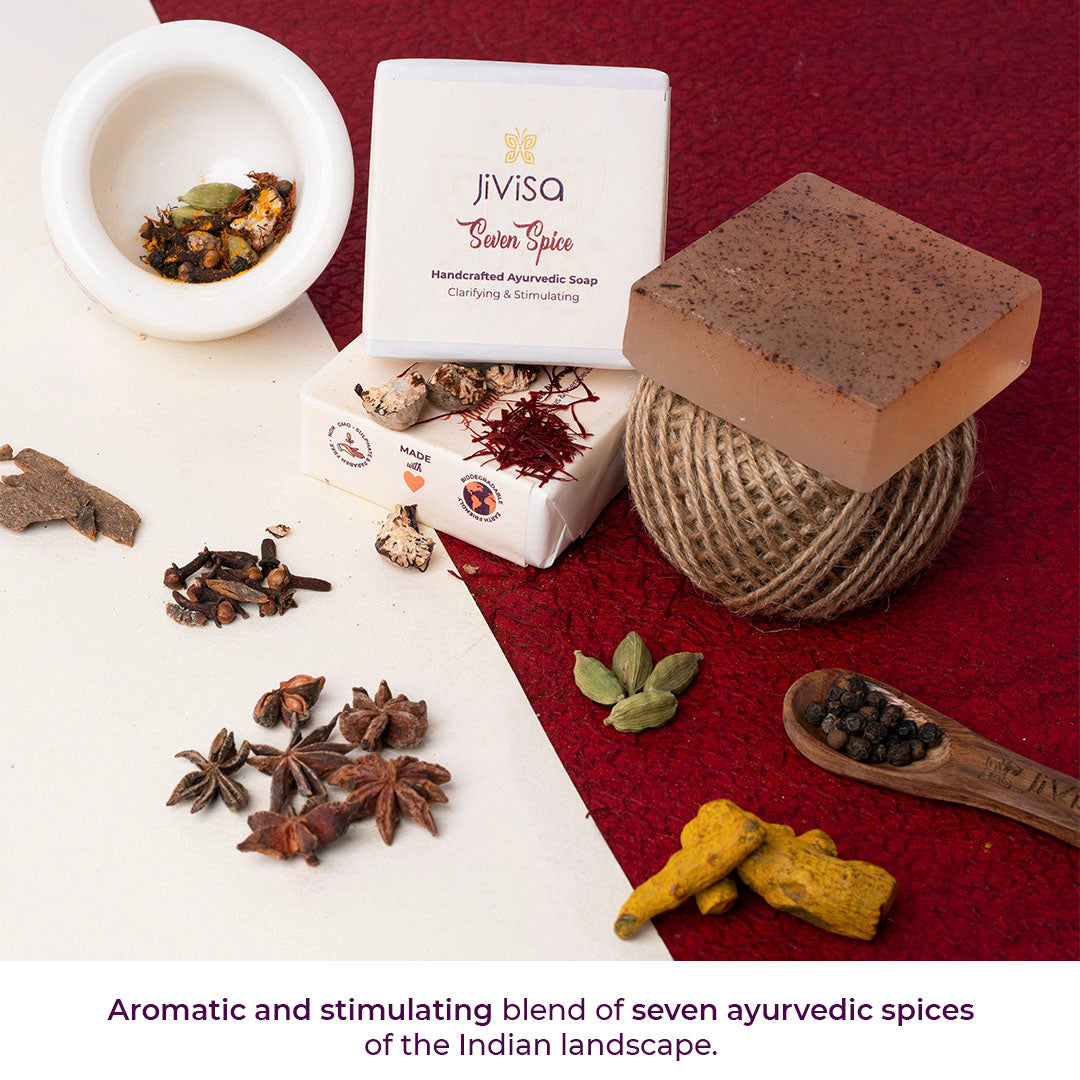An arrangement of JiViSa Seven Spice Handcrafted Ayurvedic Soap Bar, an unwrapped clear brown soap bar with visible specks and raw spices another, a white mortar with mixed spices, a ball of twine, cinnamon sticks, star anise, saffron threads, cardamom pods, and a wooden spoon with black peppercorns and turmeric roots resting on a split red and white background. Bottom text: Aromatic and stimulating blend of seven ayurvedic spices of the Indian landscape.