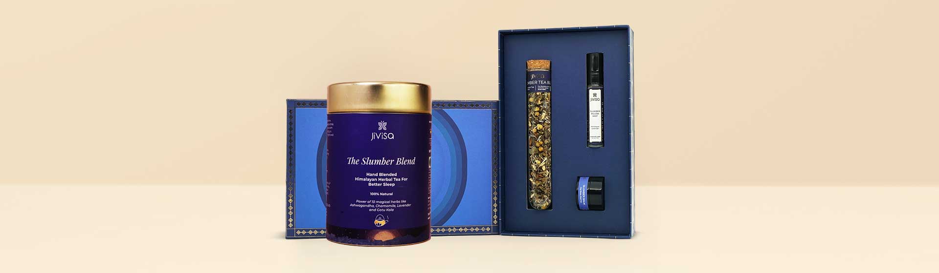 JiViSa Raina Slumber Blend gift set displayed on a beige surface, featuring calming blue and gold packaging. The box includes three products — Slumber Blend Tea in a clear glass tube filled with herbal tea leaves, Slumber Balm in a small blue jar, and Raina Slumber Pillow Mist (10ml) in a sleek spray bottle. Next to the box is a 50gm tin of JiViSa Slumber Blend Herbal Tea, crafted with soothing herbs like ashwagandha, chamomile, lavender, and gotu kola to support deep relaxation and restful sleep