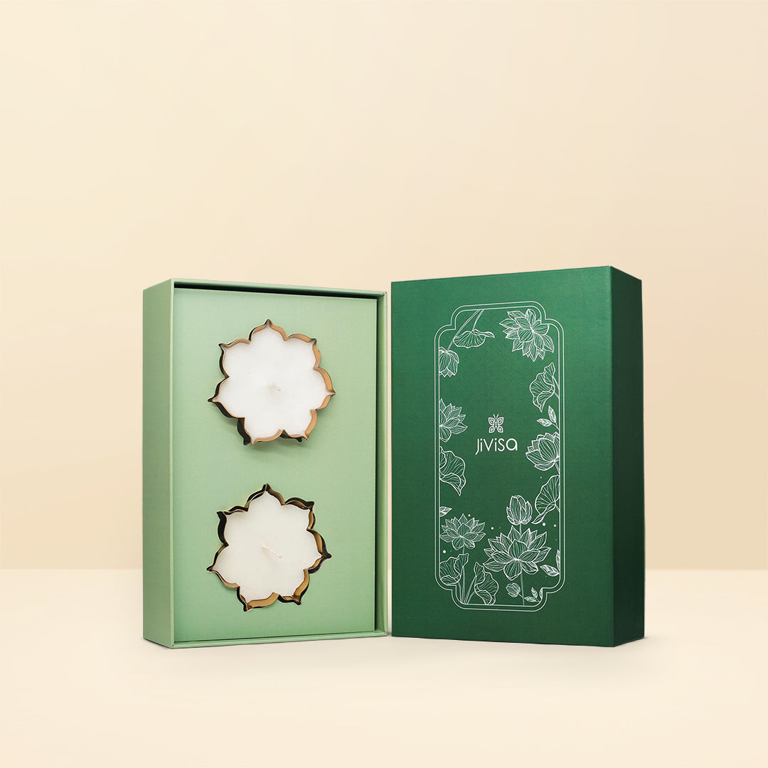 An opened JiViSa Urli Elegance Aroma gift box, reveal two white lotus-shaped candles with gold-colored edges inside a pale green insert. The deep green box lid, featuring a white outline of lotus flowers, stands up right next to the open box on a beige background.