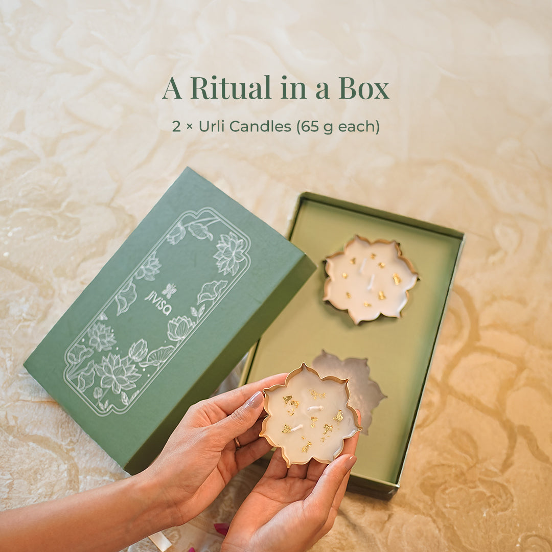 Heading reads: "A Ritual in a Box: 2 x Urli Candles (65g each)". A pair of hands gently holds one of the lotus-shaped urli candles, decorated with gold flakes, from an open JIVISA gift box. The candle&