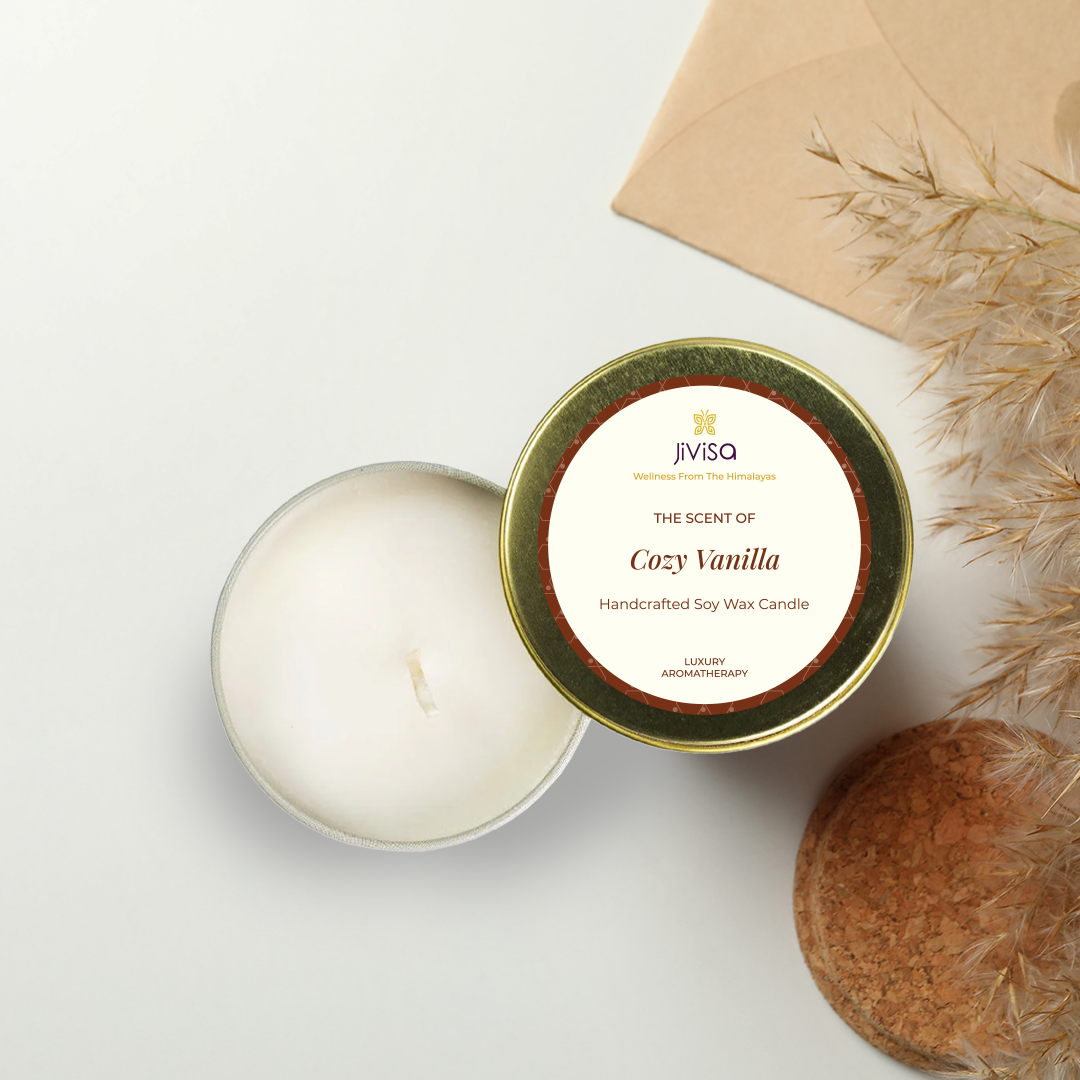 JiViSa Cozy Vanilla handcrafted soy wax candle in an open gold tin, with the lid next to it, on a white surface with a brown envelope and dried grass in the background. The label reads: "JiViSa Wellness From The Himalayas, THE SCENT OF Cozy Vanilla, Handcrafted Soy Wax Candle, LUXURY AROMATHERAPY