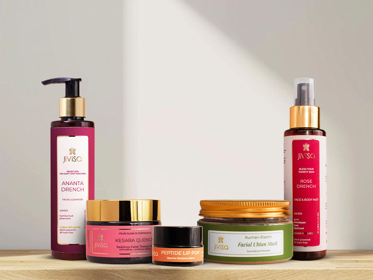 Five JiViSa clean beauty products—Ananta Drench cleanser, Kesara Quench gel moisturiser, Peptide Lip Pop, Kumari Elaichi Ubtan mask, Rose Drench face mist—on a wooden shelf, neutral background