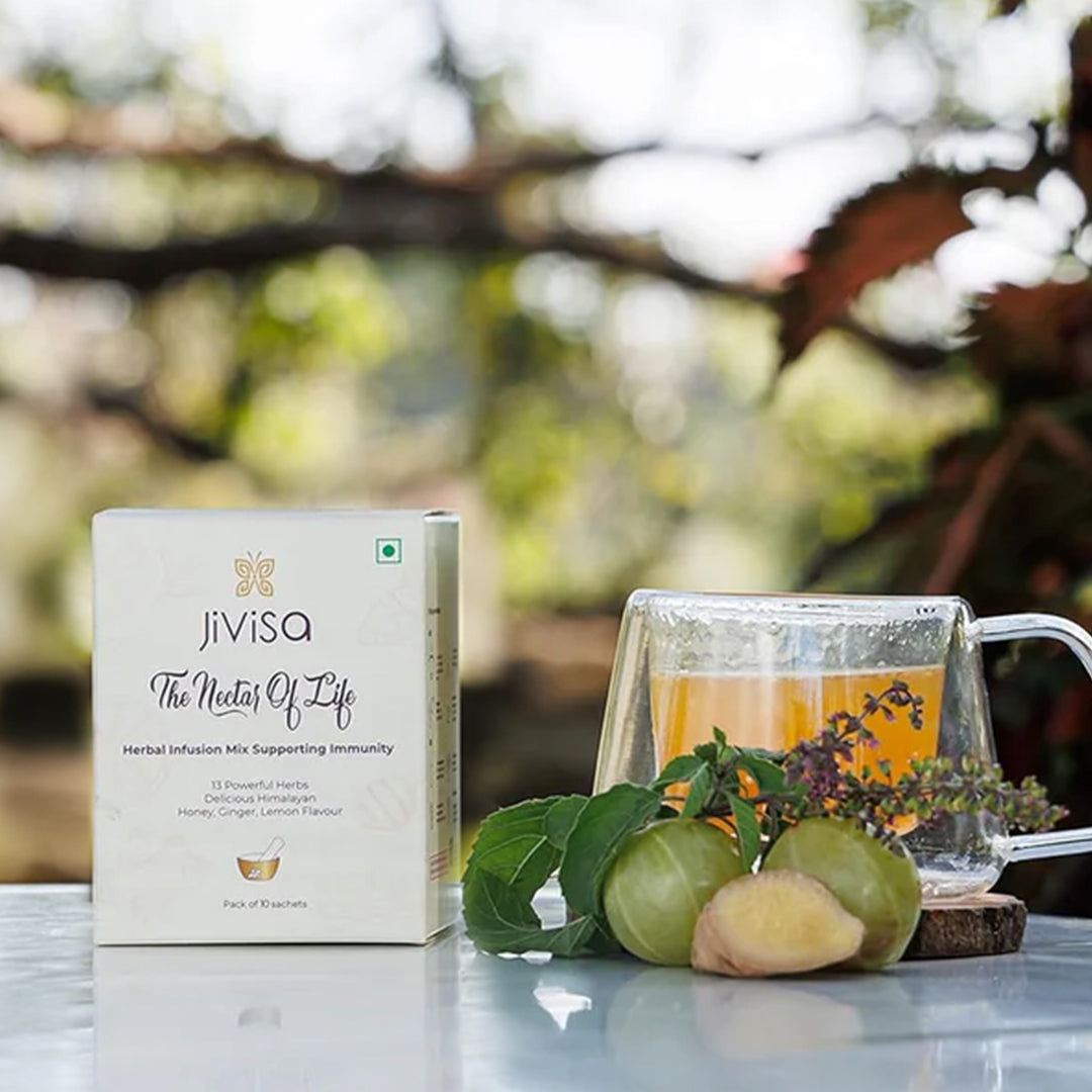 Image features a &quot;JiViSa-The Nectar of Life Herbal Infusion Mix Supporting Immunity&quot; box, indicating it contains 13 Powerful Herbs, Delicious Honey, Ginger, Lemon Flavour, and is a Pack of 10 sachets. Beside the box, a clear glass mug containing JiViSa-The Nectar of Life Herbal Infusion, along with fresh ginger, amla, and some green herbs, are arranged on a rustic wooden coaster, set against a blurred outdoor background.