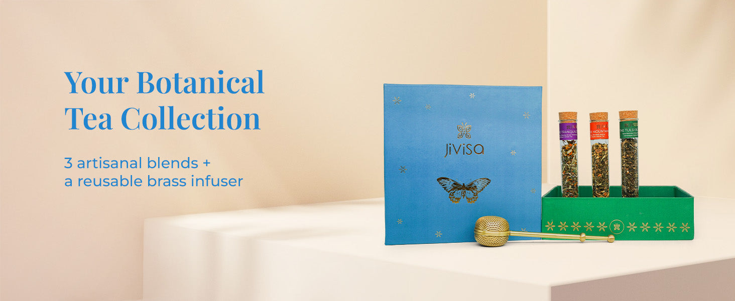 An open JiViSa Botanical Brew tea gift box placed on a white table. The box has a blue exterior with a gold butterfly and text that reads "JiViSa". Inside, a green interior holds three glass vials of JiViSa loose-leaf tea and a brass tea infuser. Tagline reads "Your Botanical Tea Collection". Below that, the text reads "3 artisanal blends + a reusable brass infuser".