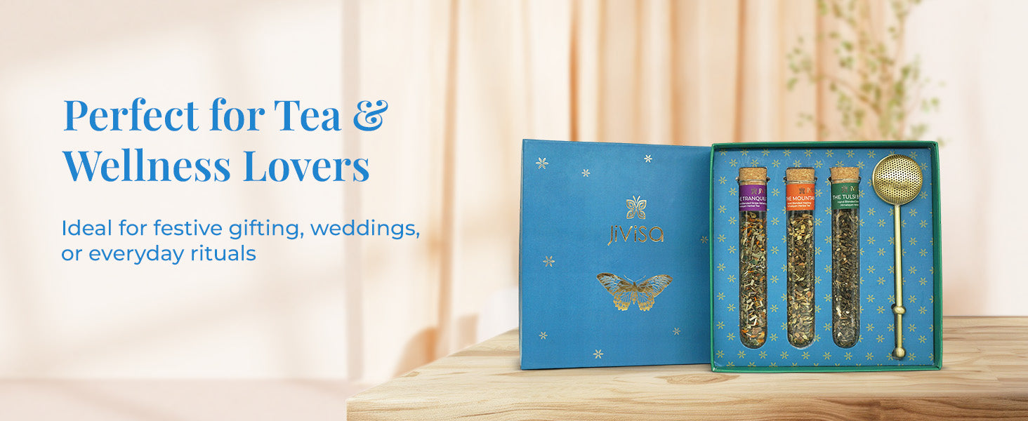 An open JiViSa Botanical Brew tea gift box placed on a wooden table. The box has a blue exterior with a gold butterfly and text that reads "JiViSa". Inside, a green interior holds three glass vials of JiViSa loose-leaf tea and a brass tea infuser. Tagline on the left reads "Perfect for Tea & Wellness Lovers" and below that, text reads: "Ideal for festive gifting, weddings, or everyday rituals".