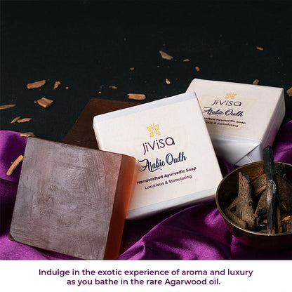 JiViSa Arabic Oudh Handcrafted Ayurvedic Soap captured in a luxurious setting with dark wood tones, rich purple silk, and natural agarwood (Oudh) pieces in a copper bowl. An unwrapped soap bar with a deep brown translucent tone is also displayed, emphasizing its natural composition. Below the setup, the text reads: “Indulge in the exotic experience of aroma and luxury as you bath in the rare Agarwood Oil&quot;.
