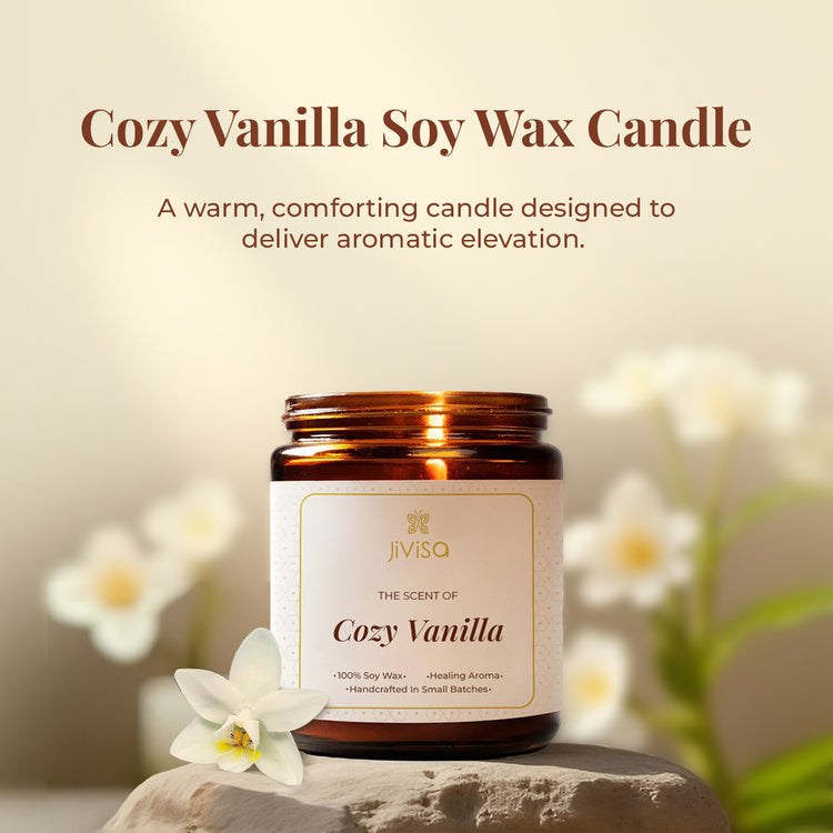  JiViSa Cozy Vanilla candle placed on a stone surface with white vanilla flowers in the background. The tagline above reads: "Cozy Vanilla Soy Wax Candle – A warm, comforting candle designed to deliver aromatic elevation." The label on the jar reads: "The Scent of Cozy Vanilla – 100% Soy Wax • Healing Aroma • Handcrafted In Small Batches."