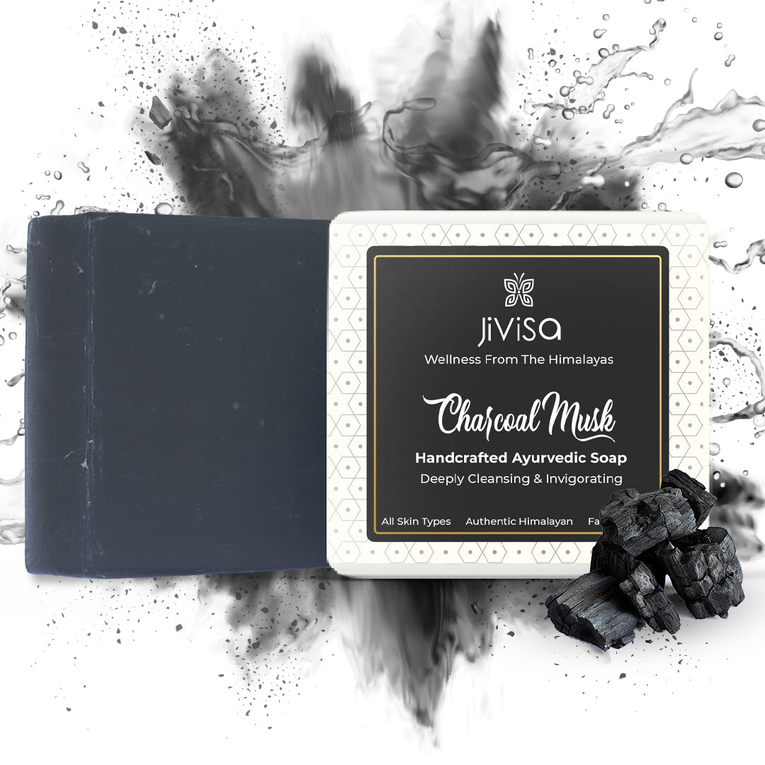 JiViSa Charcoal Musk Handcrafted Ayurvedic Black Colored Soap bar, next to its white and black packaged box.  A pile of charcoal pieces is in the foreground. The background is white with a dynamic black splash and water droplet effect, suggesting .cleansing