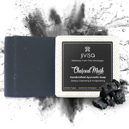 JiViSa Charcoal Musk Handcrafted Ayurvedic Black Colored Soap bar, next to its white and black packaged box.  A pile of charcoal pieces is in the foreground. The background is white with a dynamic black splash and water droplet effect, suggesting .cleansing
