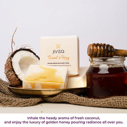 JiViSa Coconut &amp; Honey Handcrafted Ayurvedic Soap bar placed next to its white and gold packaged box. A broken coconut half, a glass jar of honey with a dipper are arranged around it on a textured surface. Bottom text reads: Inhale the heady aroma of fresh coconut and enjoy the luxury of golden honey pouring radiance all over you.