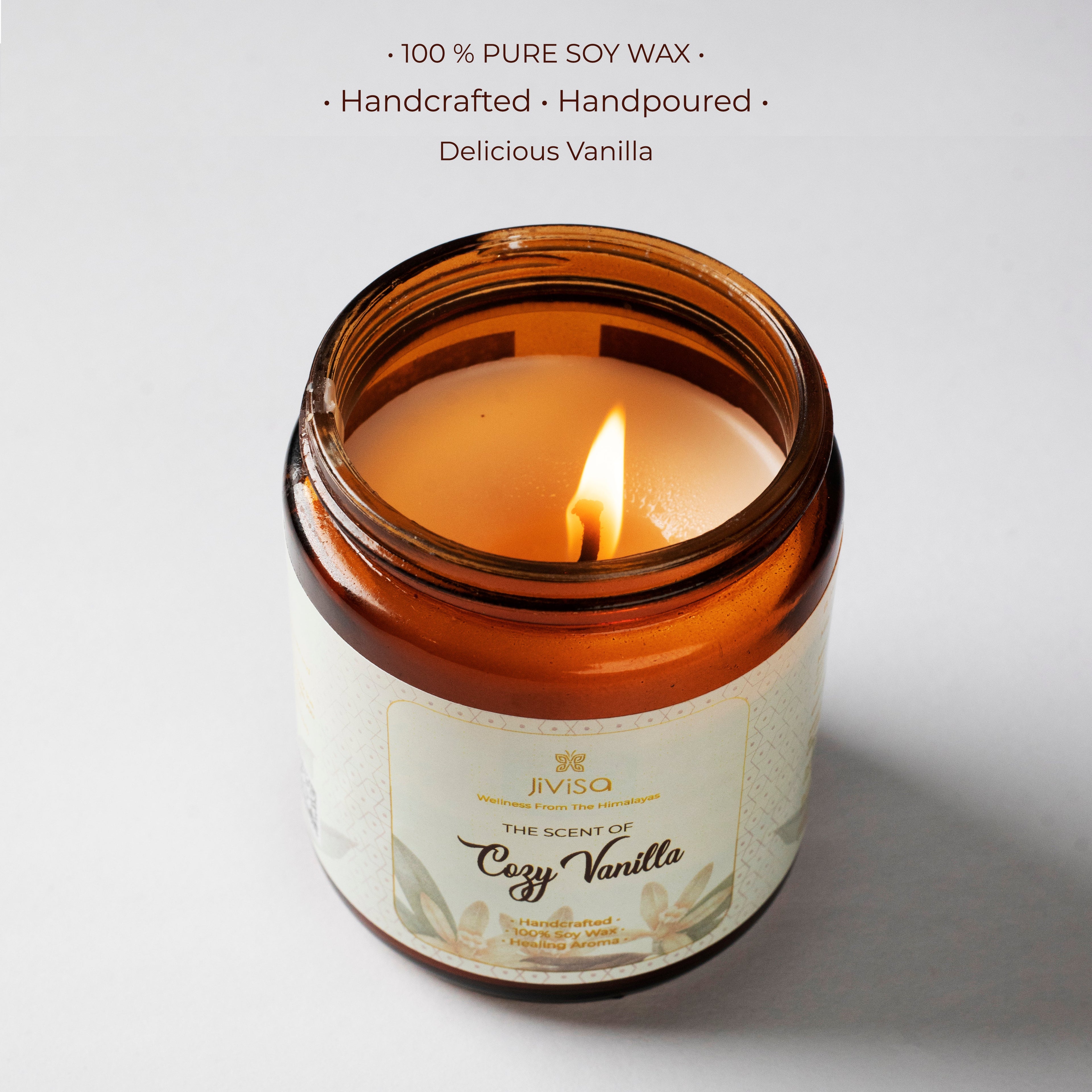 A top-down view of the lit JiViSa Cozy Vanilla candle, showcasing the smooth soy wax surface with a warm amber flame. The jar is amber glass with a detailed label reading &quot;The Scent of Cozy Vanilla.&quot; Above the image, text reads: &quot;100% PURE SOY WAX • Handcrafted • Handpoured • Delicious Vanilla,&quot; highlighting the purity and artisanal quality of the product.
