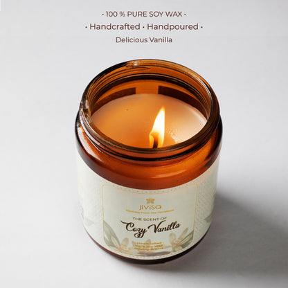A top-down view of the lit JiViSa Cozy Vanilla candle, showcasing the smooth soy wax surface with a warm amber flame. The jar is amber glass with a detailed label reading &quot;The Scent of Cozy Vanilla.&quot; Above the image, text reads: &quot;100% PURE SOY WAX • Handcrafted • Handpoured • Delicious Vanilla,&quot; highlighting the purity and artisanal quality of the product.