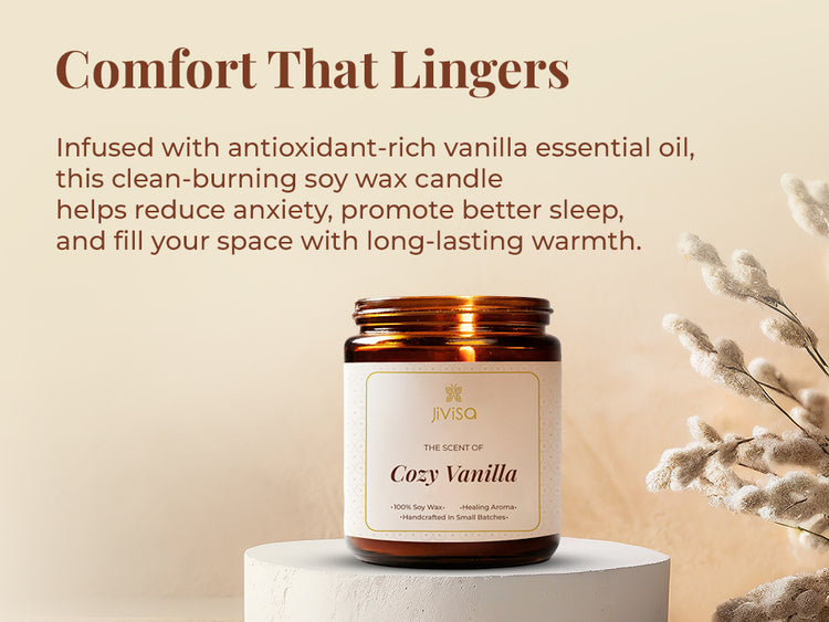 JiViSa Cozy Vanilla soy wax candle glass jar is displayed on a light pedestal against a beige, softly-lit backdrop with dried foliage accents, creating a warm and calming atmosphere. The tagline reads: "Comfort That Lingers," followed by the text: "Infused with antioxidant-rich vanilla essential oil, this clean-burning soy wax candle helps reduce anxiety, promote better sleep, and fill your space with long-lasting warmth."