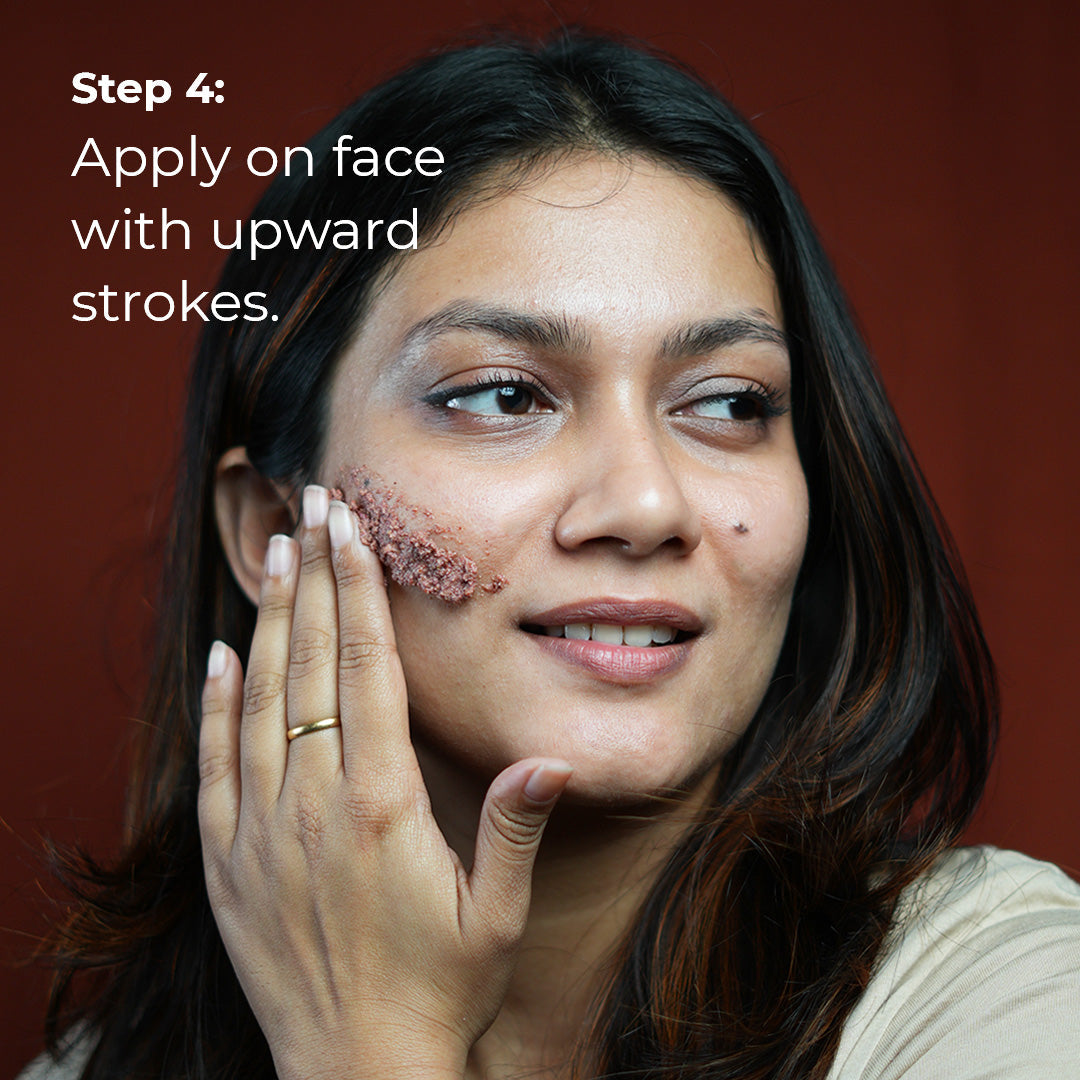 A woman with medium brown skin smiles gently as she applies a light purple scrub to her cheek with upward strokes. She is wearing a simple gold ring. Text overlay reads &quot;Step 4: Apply on face with upward strokes