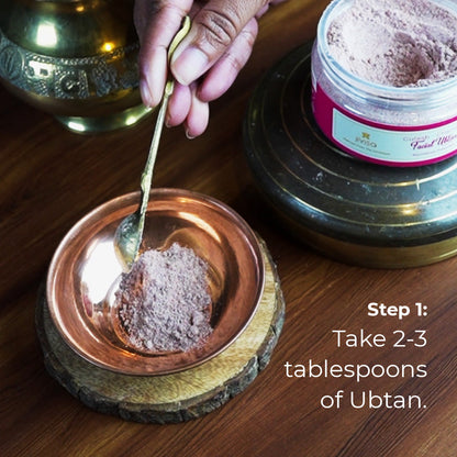 A hand holds a golden spoon scooping a generous amount of light brown, finely textured powder, identified as JiViSa Gulaab Chandan Facial Ubtan. The powder is being transferred into a small copper bowl that sits on a circular, light-colored wooden coaster. The text on the image highlights the instruction:
 Step 1: Take 2-3 tablespoons of Ubtan.