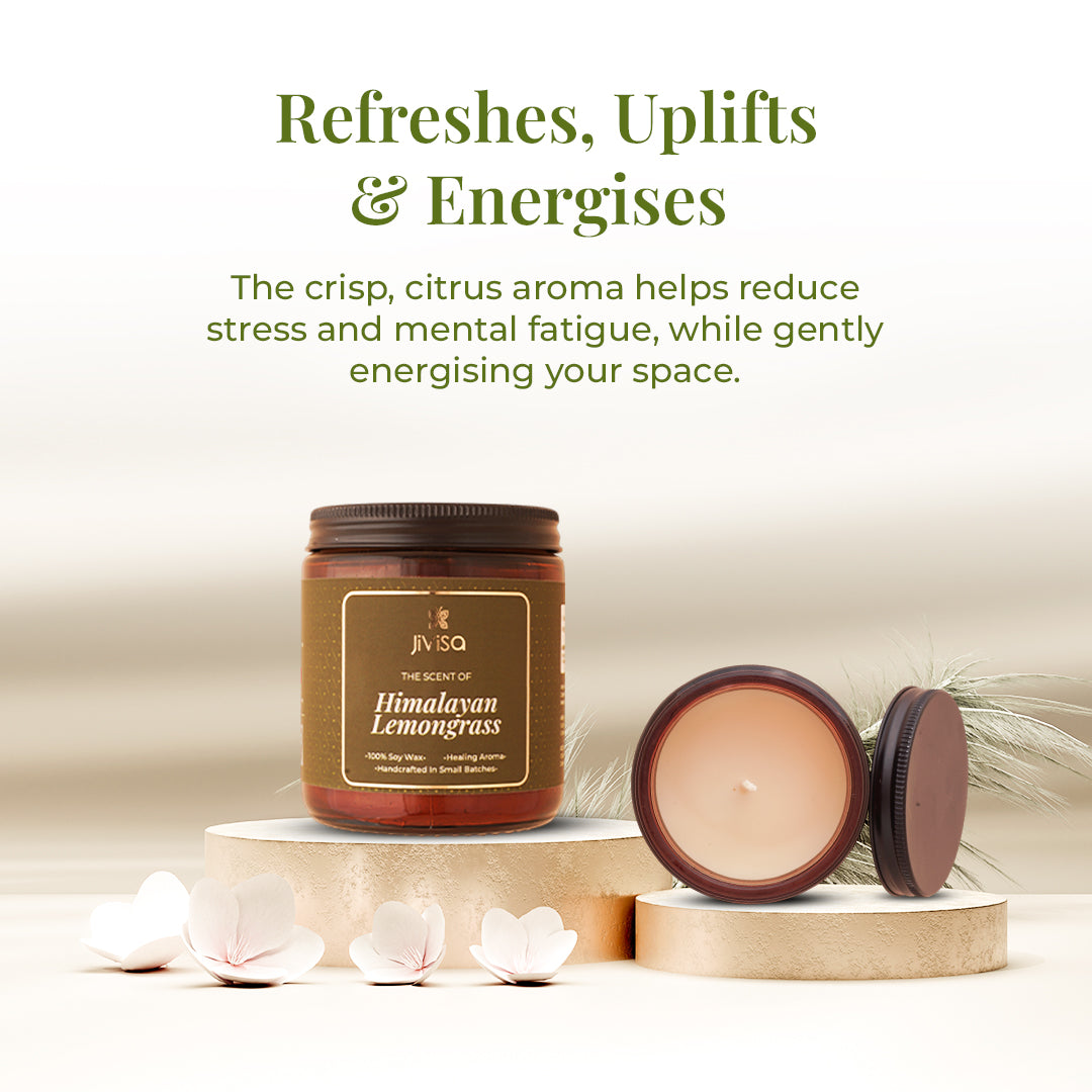 A styled display of the JiViSa Himalayan Lemongrass soy wax candle glass jar alongside an open lid and soft floral elements. The headline above reads: "Refreshes, Uplifts &amp; Energises." Supporting text says: "The crisp, citrus aroma helps reduce stress and mental fatigue, while gently energising your space."

