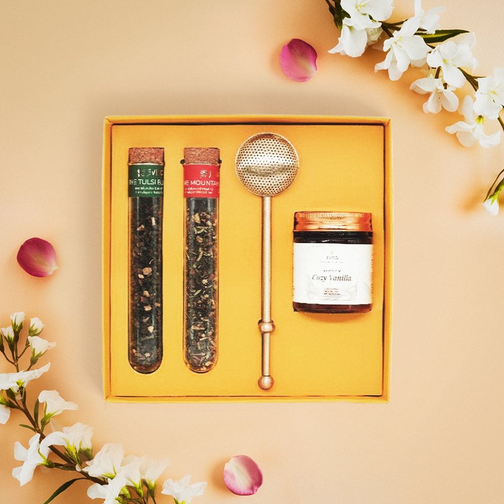 JiViSa Brew &amp; Bliss Gift Set contain two clear glass tubes of JiViSa- The Tulsi Blend and JiViSa- The Mountain Blend each sealed with a cork stopper and containing loose-leaf tea, a gold-toned, long-handled tea infuser with a mesh ball at the end, designed for brewing loose-leaf tea, and also a JiViSa-Cozy Vanilla soy wax candle. The Gift presented in a bright yellow box arranged on a light peach-colored surface adorned with scattered pink petals and sprigs of white flowers.
