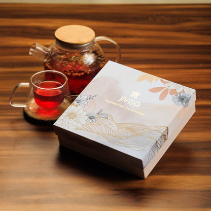 JiViSa Himalayan Discovery Tea Gift Box featuring the tagline &quot;Wellness From The Himalayas&quot; on the lid. Below the gift box, clear glass tea pot and a cup are positioned  with read brown herbal tea.