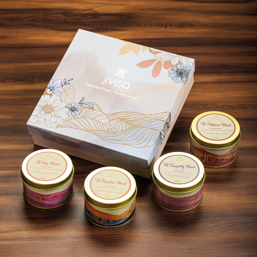 JiViSa Himalayan Discovery Tea Gift Box. A box decorated with delicate floral line art in gold and silver, along with the &quot;JiViSa&quot; logo and the tagline Wellness From The Himalayas. Below the box are four round tins of JiViSa premium loose-leaf tea. From left to right, the tins are labeled The Rose Blend with pink and red watercolor-style, The Mountain Blend with blue and orange watercolor-style, The Tranquility Blend with purple watercolor-style label and The Hibiscus with red style label.