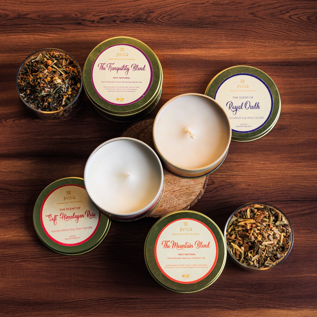JiViSa Premium Loose Leaf Tea and Soy wax Candle Gift Box. The image displays a selection of JiViSa products arranged on a dark wooden surface. There are four round tins, two of which appear to contain tea (&quot;The Tranquility Blend&quot; and &quot;The Mountain Blend,&quot; both labeled &quot;100% Natural&quot;), and two appear to be soy wax candles (&quot;The Scent of Soft Himalayan Rose&quot; and &quot;The Scent of Royal Oudh&quot;)