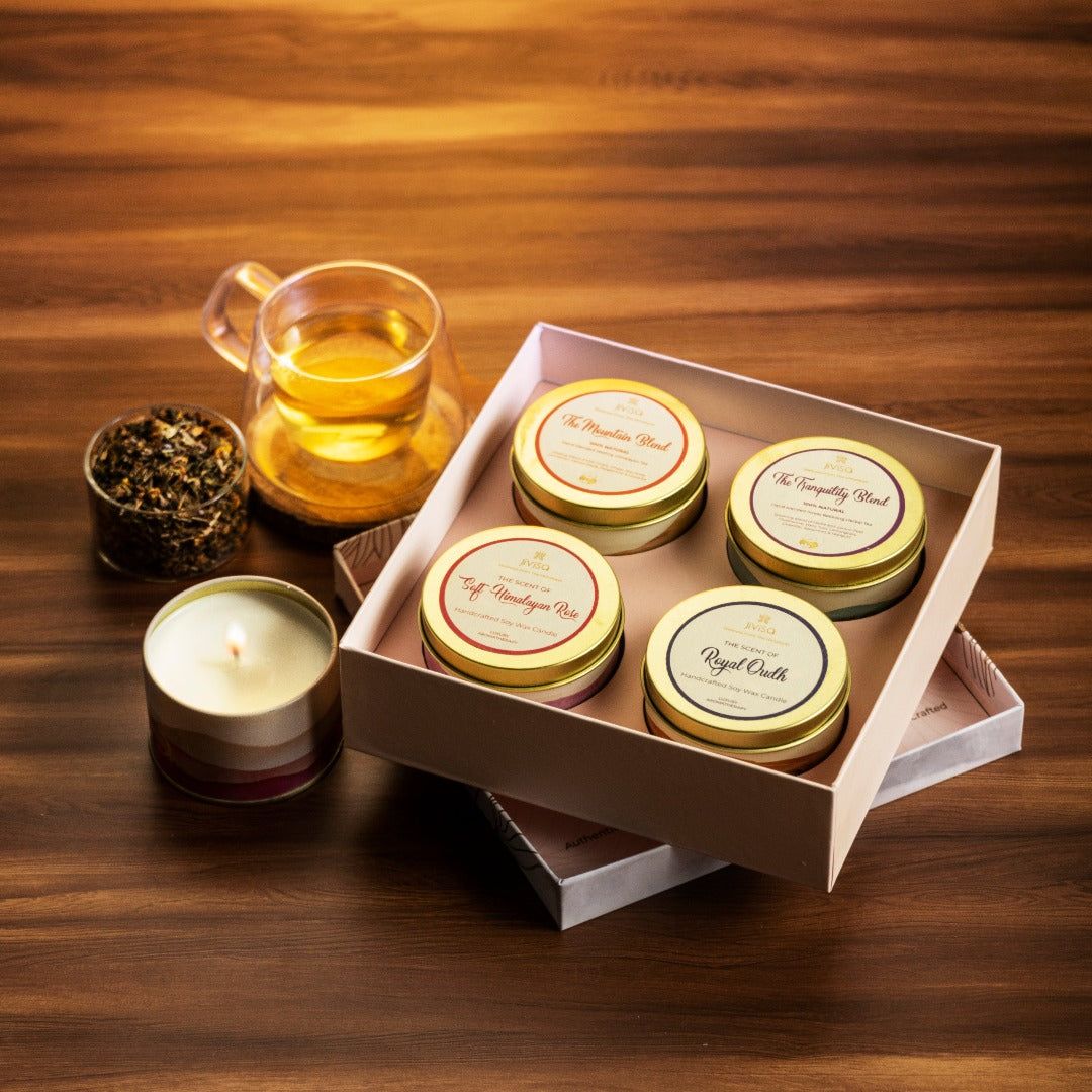JiViSa Premium Loose Leaf Tea and Soy wax Candle Gift Box. The box contains four round tins arranged: The teas are &quot;The Mountain Blend,&quot; and &quot;The Tranquility Blend,&quot; and Soy wax Candle are &quot;Soft Himalayan Rose&quot; and &quot;Royal Oudh&quot;. Next to the gift box is a clear glass cup with a light-colored beverage, a clear glass cup containing tea leaves and a small, round candle.