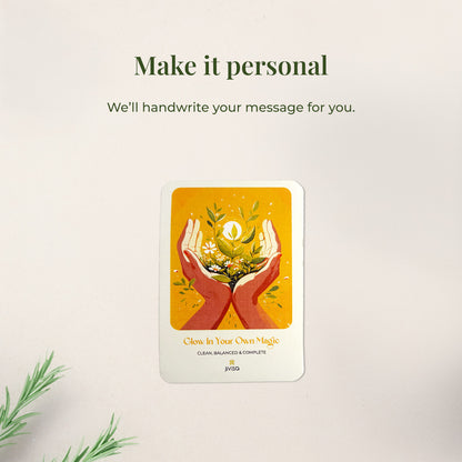 &quot;Make it personal – We’ll handwrite your message for you.&quot; A handwritten card with warm yellow and orange tones shows two hands holding blooming herbs with a glowing sun. Placed on a white surface, the image emphasizes the personal, heartfelt gifting experience JiViSa offers.