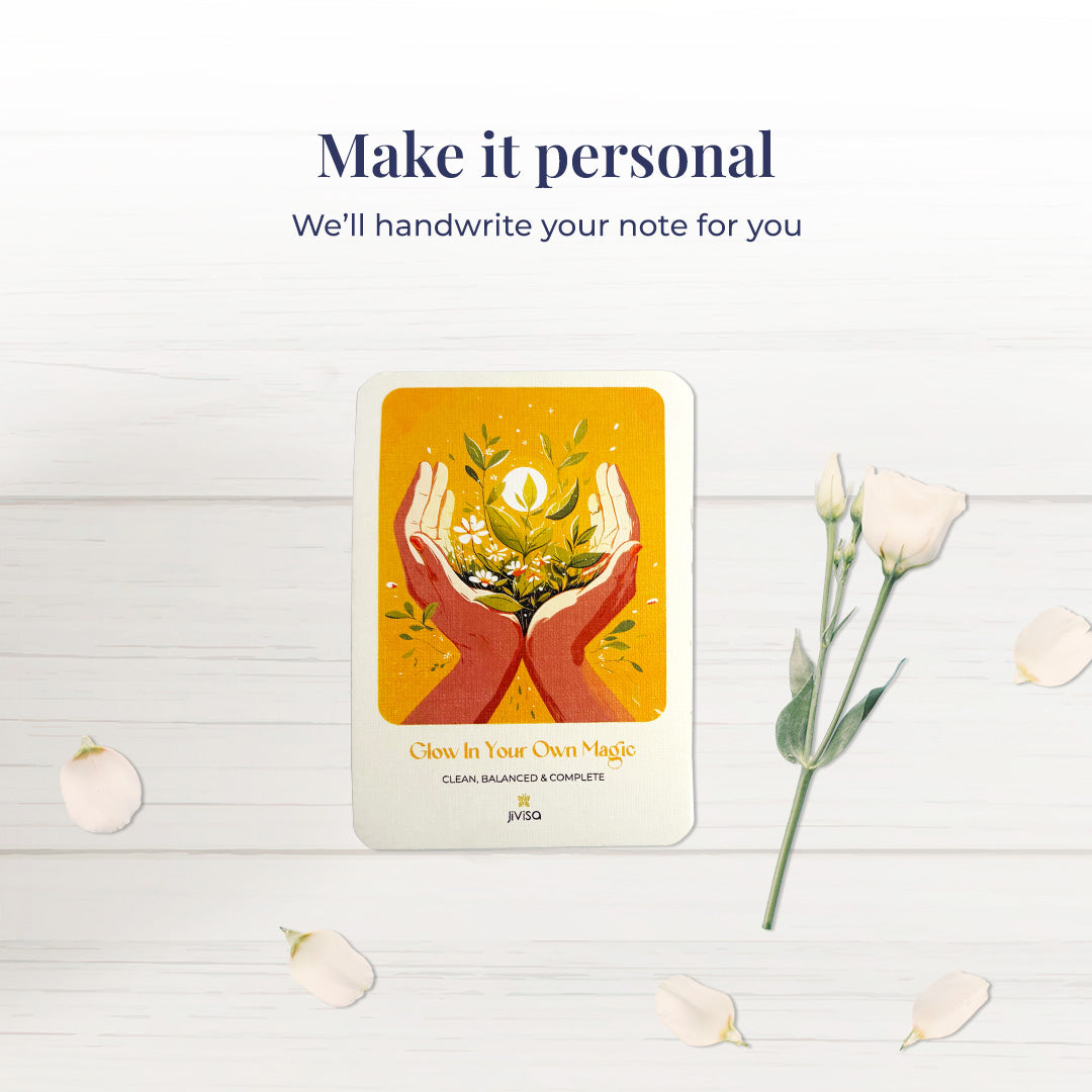 &quot;Make it personal – We’ll handwrite your message for you.&quot; A handwritten card with warm yellow and orange tones shows two hands holding blooming herbs with a glowing sun. Placed on a white surface with scattered white rose petals and a white bloom, emphasizing the personalized gifting experience.