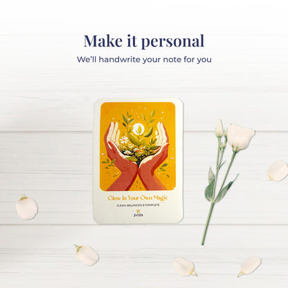 &quot;Make it personal – We’ll handwrite your message for you.&quot; A handwritten card with warm yellow and orange tones shows two hands holding blooming herbs with a glowing sun. Placed on a white surface with scattered white rose petals and a white bloom, emphasizing the personalized gifting experience.
