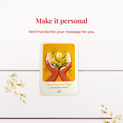 &quot;Make it personal – We’ll handwrite your message for you.&quot; A handwritten card with warm yellow and orange tones shows two hands holding blooming herbs with a glowing sun. Placed on a white wooden surface with jasmine flowers scattered around, the image emphasizes the personal, heartfelt gifting experience JiViSa offers.