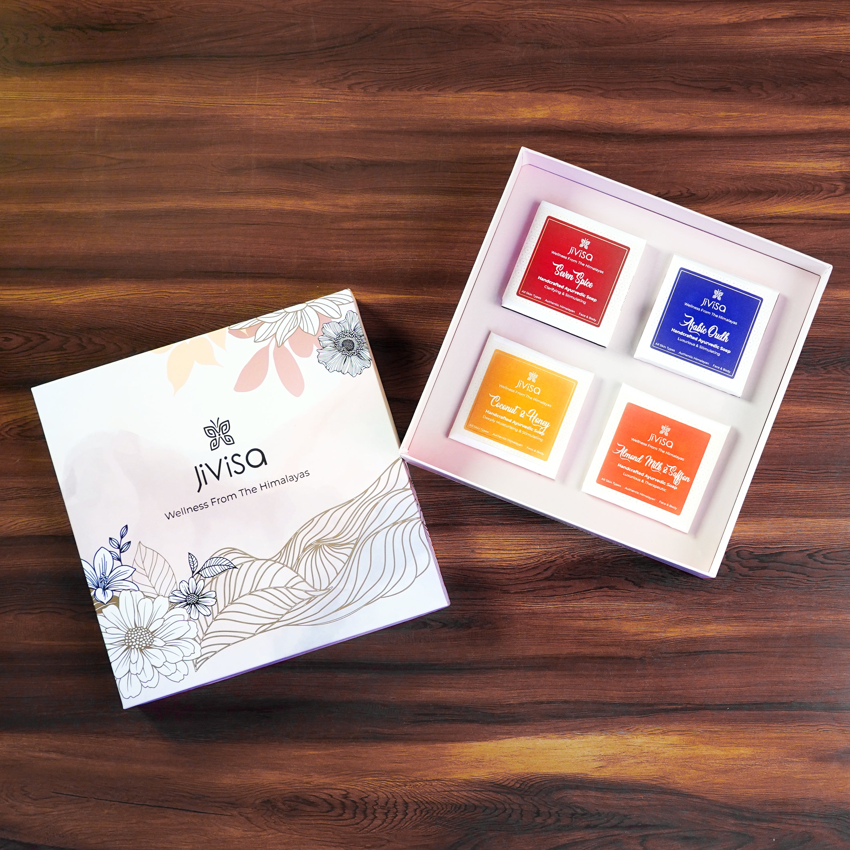 JiViSa-Ayurvedic Glow Soap Gift Box with four handcrafted soaps in floral-themed packaging, displayed on a wooden surface. JiViSa-Ayurvedic Glow Soap Gift Box contains Seven Spice Ayurvedic Soap – a revitalizing blend inspired by ancient Indian spice therapy, Arabic Oudh Ayurvedic Soap – a deep, aromatic experience with warm, woody notes, Coconut &amp; Honey Ayurvedic Soap– nourishes and softens skin, Almond Milk &amp; Saffron Ayurvedic Soap– a skin-brightening and soothing bar enriched with royal Ayurvedic herbs