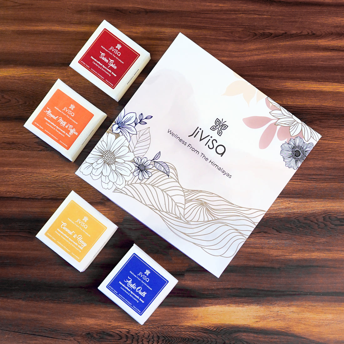 JiViSa Luxury Ayurvedic Handmade Soaps Gift Box. JiViSa four distinct Handmade soaps: Seven Spices, Arabic Oudh, Coconut &amp; Honey and Almond Milk &amp; Saffron are placed on the wooden surface around the box.